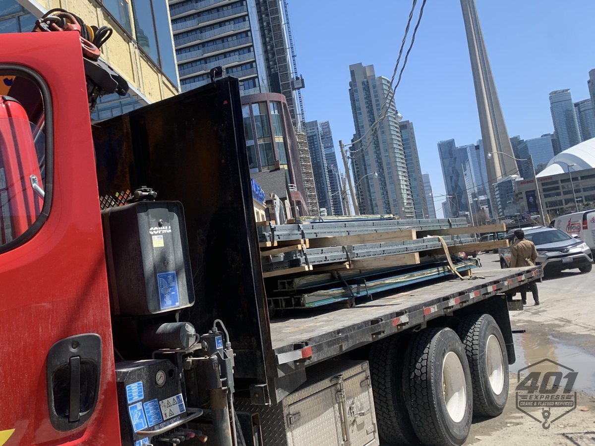 401 Express flatbed loaded with elevator guide rails at a downtown Toronto high-rise site with the CN Tower behind