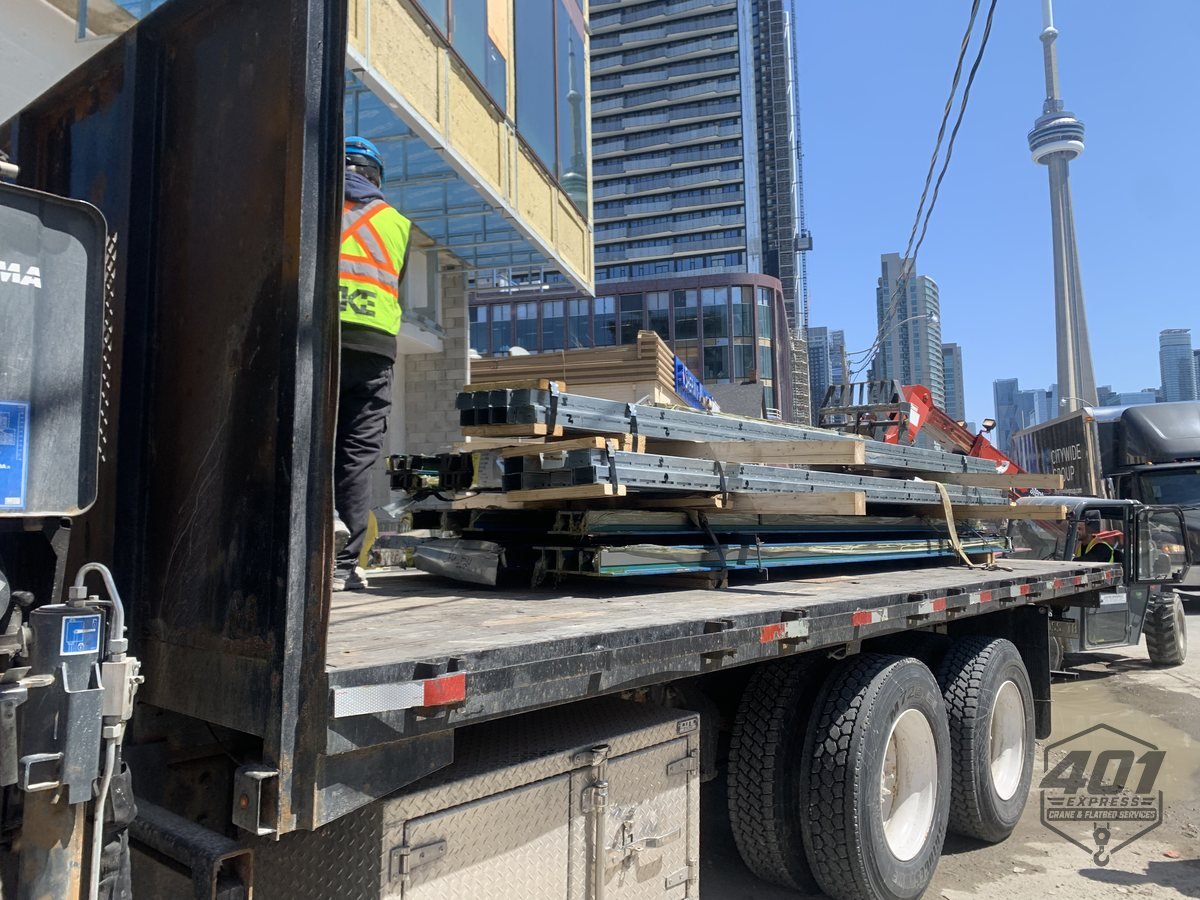 Elevator guide rails being unloaded from the 401 Express flatbed by a worker in full PPE at a downtown Toronto high-rise, CN Tower behind